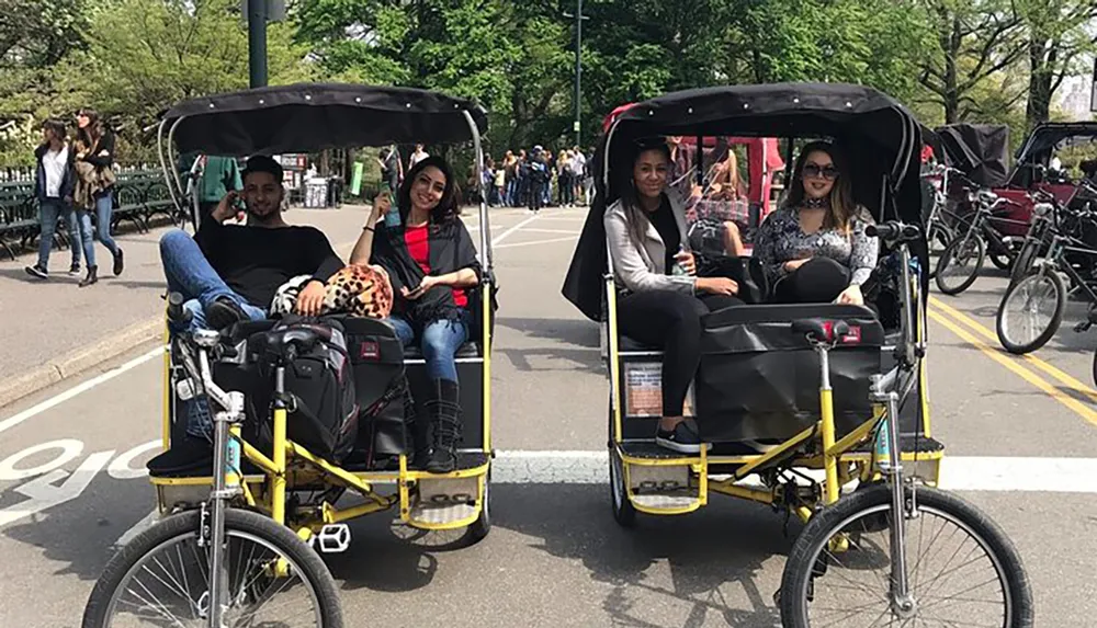 Two pedicabs carry smiling passengers on what appears to be a pleasant day in a busy park or urban area