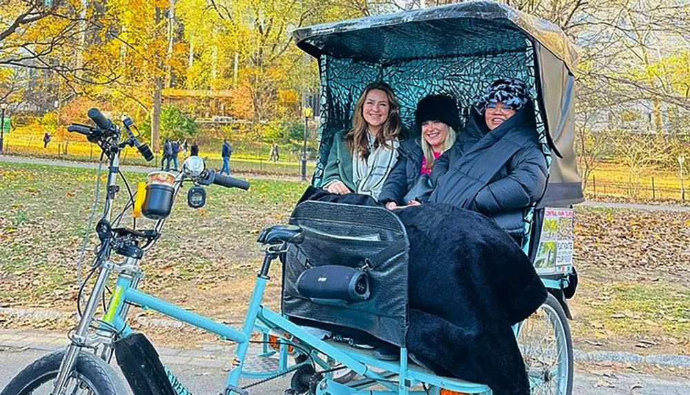 Three people are smiling while sitting in a pedicab in a park with autumn foliage in the background