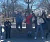 A group of people is posing joyfully with raised arms in front of a fountain in a park on a sunny day