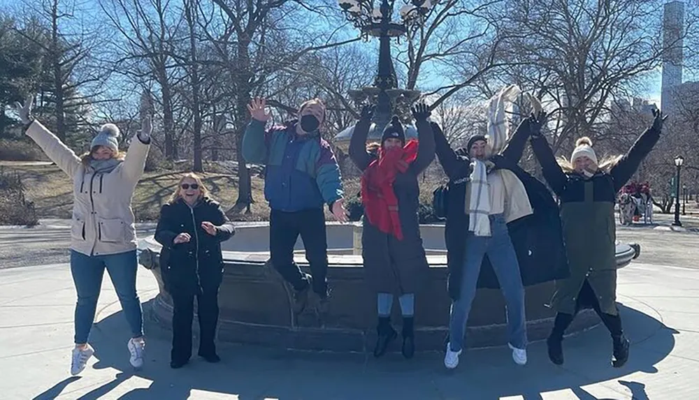 A group of people is posing joyfully with raised arms in front of a fountain in a park on a sunny day