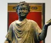 This image shows a bronze sculpture of a young boy in classical style raising his right hand possibly in gesture with a smiling expression and curly hair set against a blurred background with red and black elements