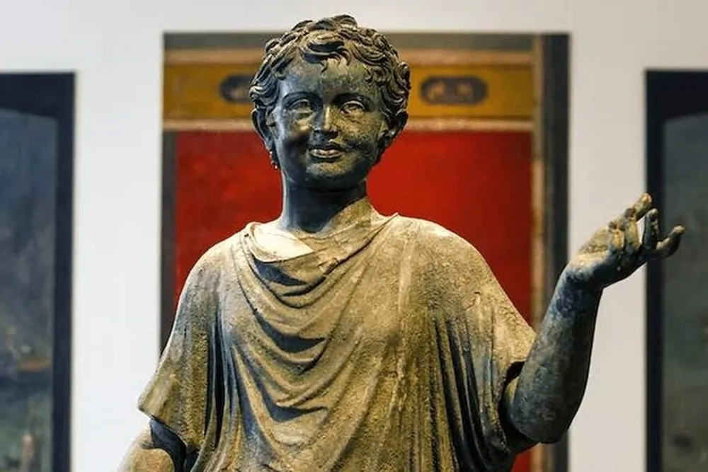 This image shows a bronze sculpture of a young boy in classical style raising his right hand possibly in gesture with a smiling expression and curly hair set against a blurred background with red and black elements