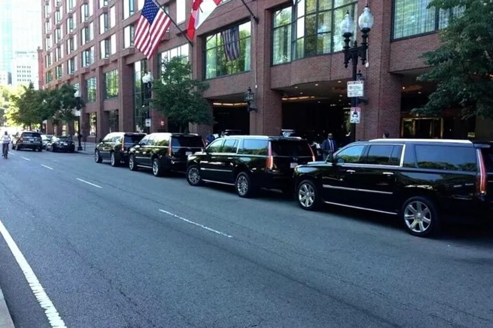 A motorcade of black SUVs is lined up in front of a building with a row of flags hanging above