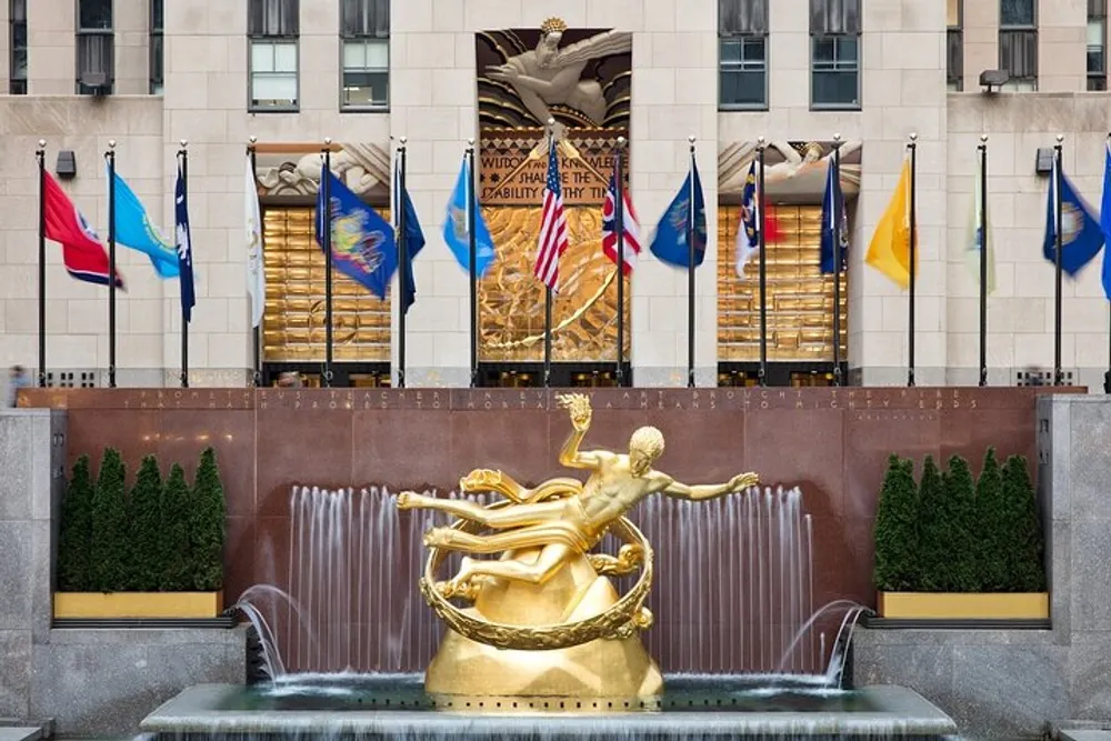 The image shows the iconic golden Prometheus statue overlooking the ice skating rink at Rockefeller Center against a backdrop of international flags and the bold artwork above the rink