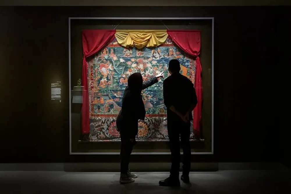 Two people are observing and discussing a colorful tapestry displayed in a dimly lit museum gallery