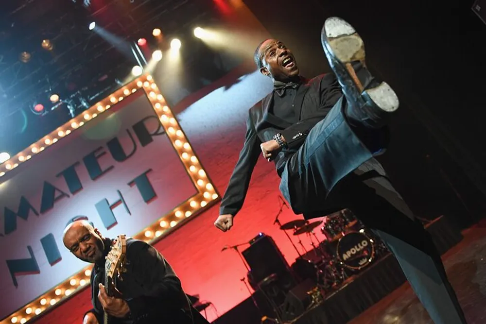 A performer is kicking high in the air while another plays the guitar during an exuberant stage performance with Amateur Night displayed in the background