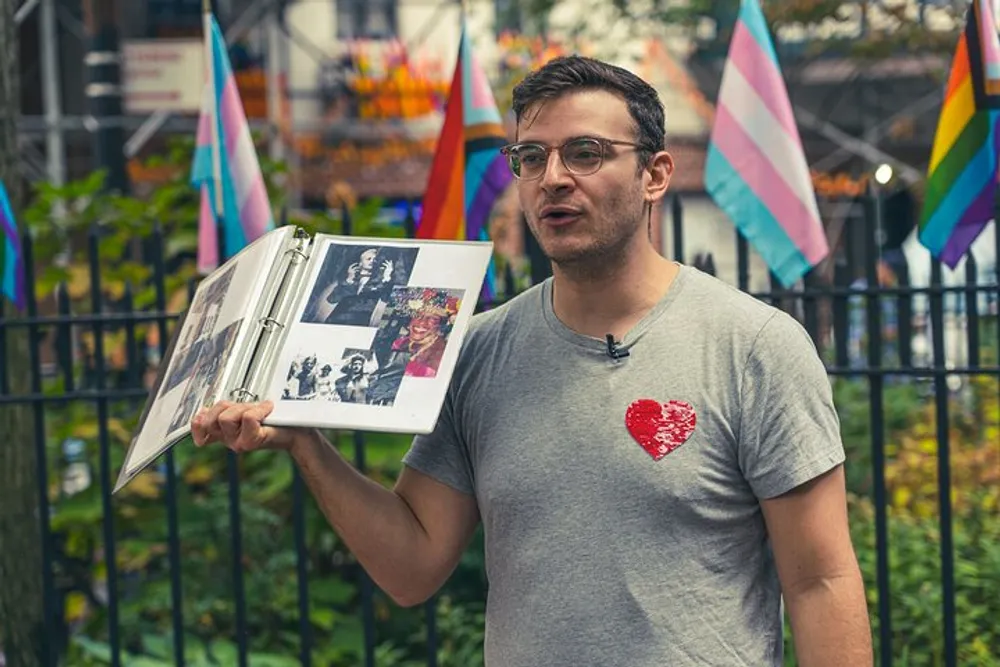 A person is holding an open photo album while standing in front of a fence adorned with colorful pride flags