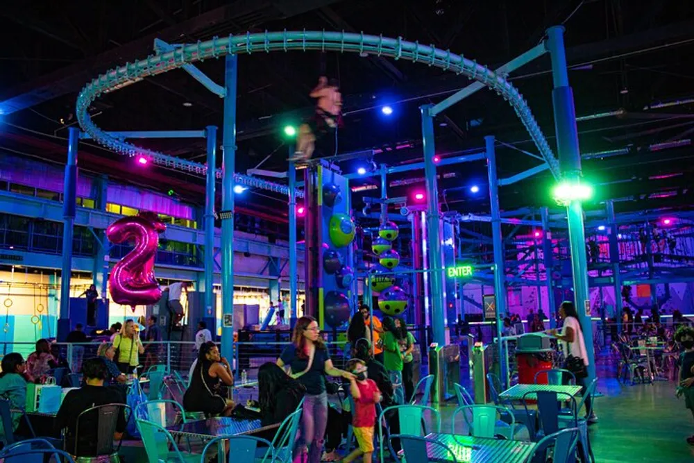 The image depicts an indoor amusement center with vibrant lighting featuring a roller coaster track above various play structures and people seated and walking around