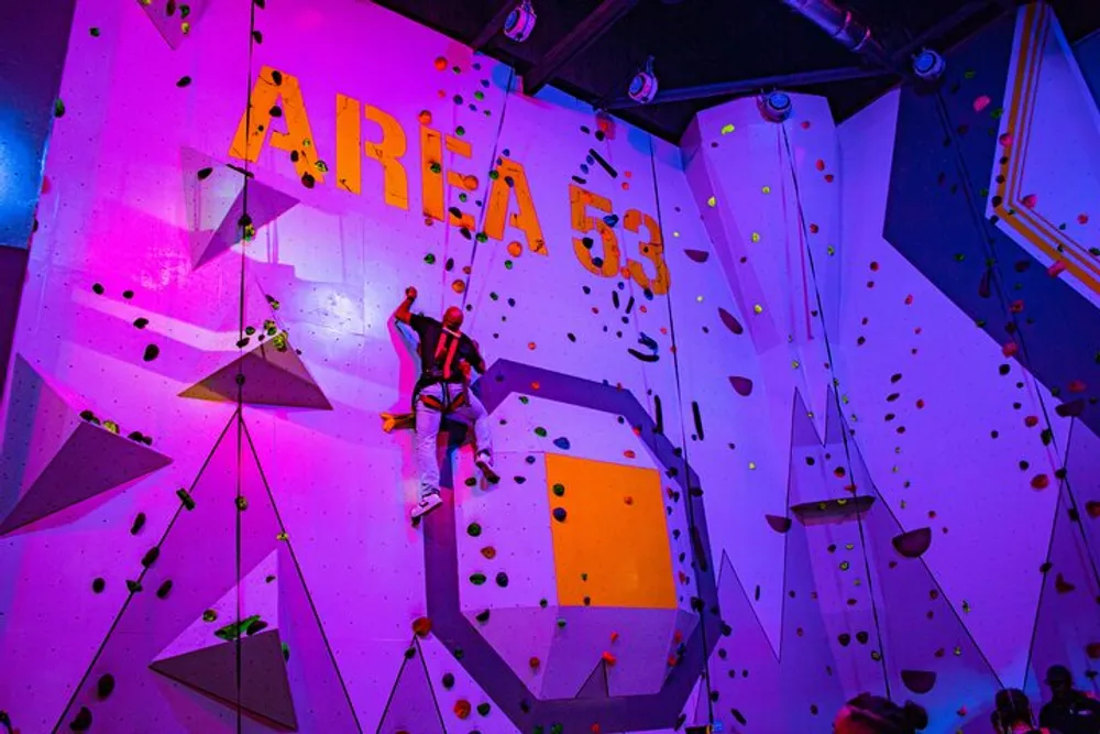 A person is climbing an indoor rock wall labeled AREA 55 with various colorful holds and geometric shapes under vibrant purple lighting