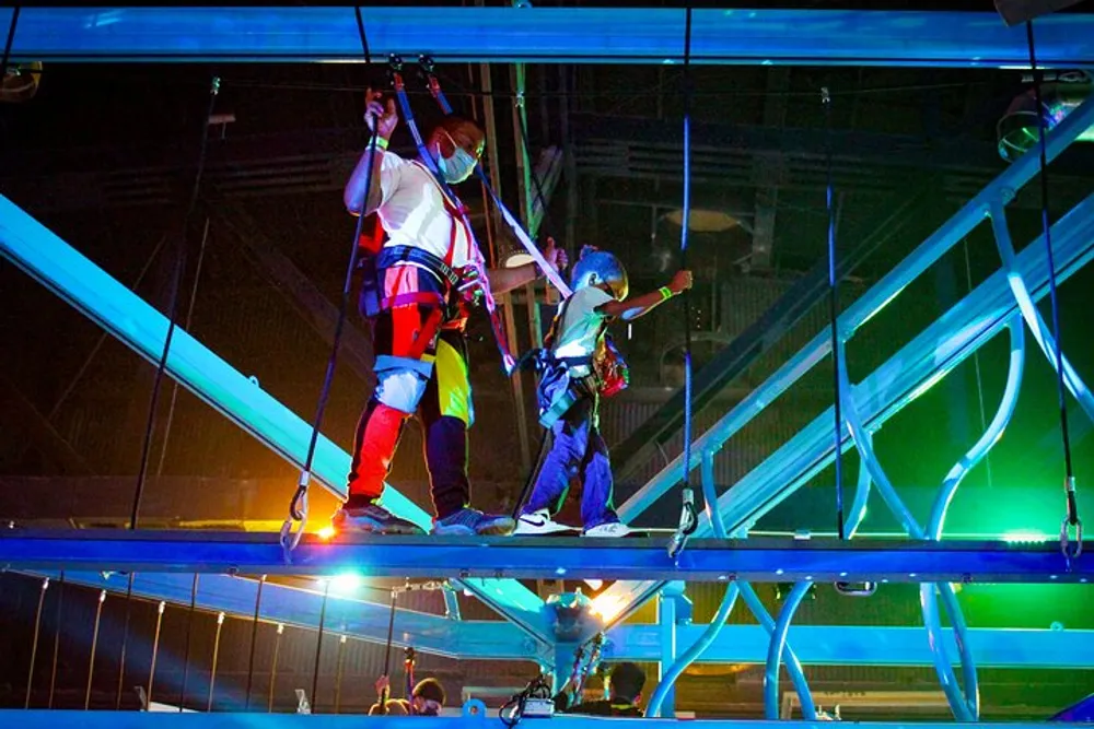 Two individuals wearing safety harnesses are navigating a high indoor rope course amidst a network of metal beams illuminated by colorful lights