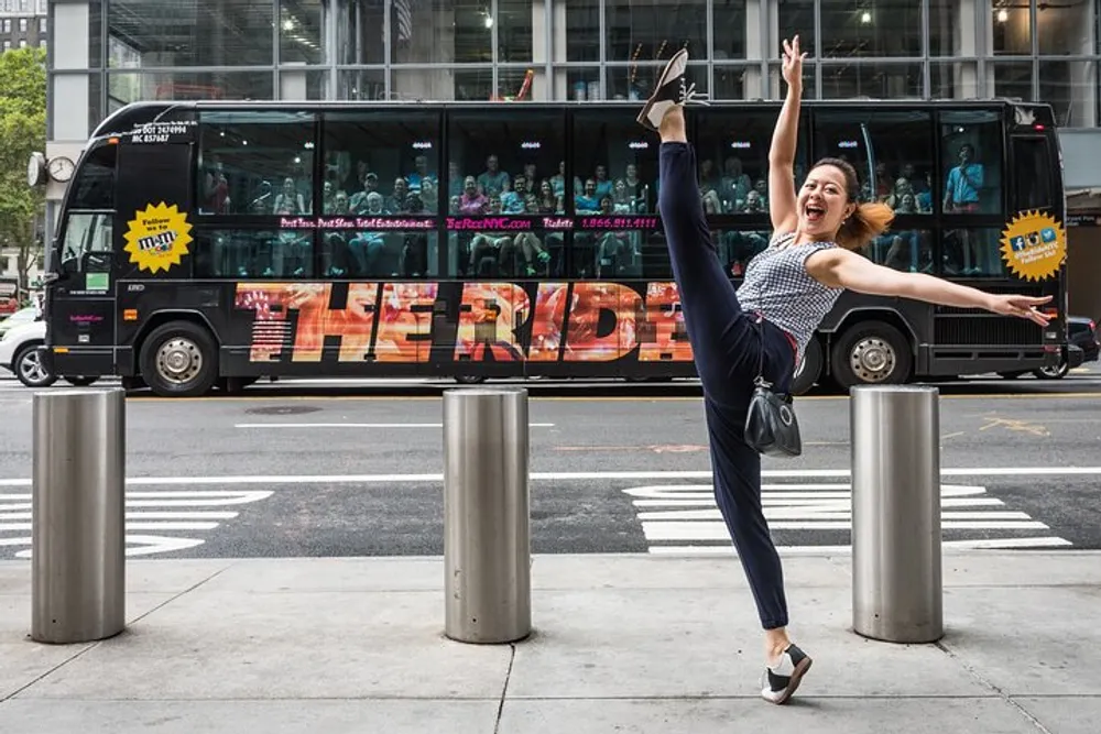 A joyful woman is striking a playful dance pose on the sidewalk with a bus labeled THE RIDE in the background