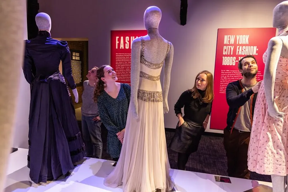 Visitors are admiring a display of vintage dresses at a fashion exhibition called New York City Fashion 1880s-1960s