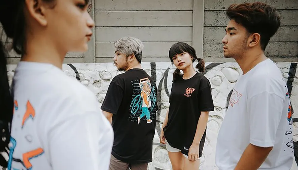 Four young adults are casually dressed in graphic tees standing with a neutral demeanor against a concrete backdrop