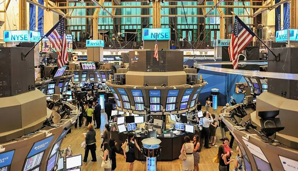 The image shows the bustling floor of the New York Stock Exchange with traders working at their desks surrounded by electronic screens and flags