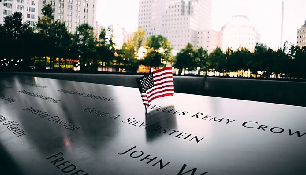 The image features an American flag placed beside names engraved on a reflective memorial with trees and buildings softly illuminated in the background at dusk