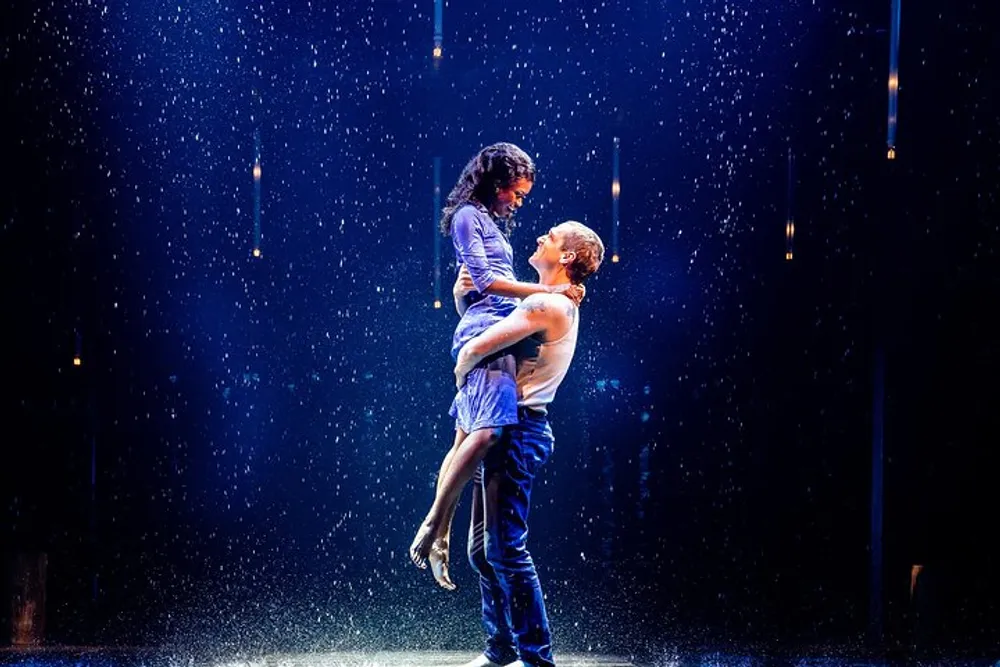 A man lifts a woman in a graceful pose on a stage with illuminated rain-like effects possibly during a dance performance or a theatrical act