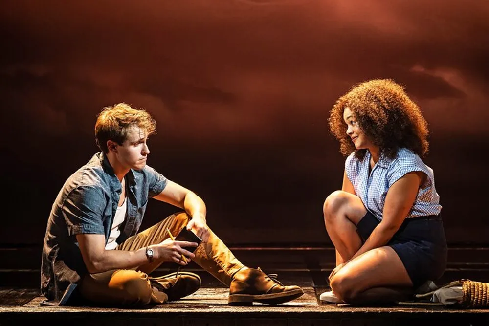 Two actors are sitting on a stage engaging with each other under dramatic lighting that suggests an intimate or significant moment in a theatrical performance