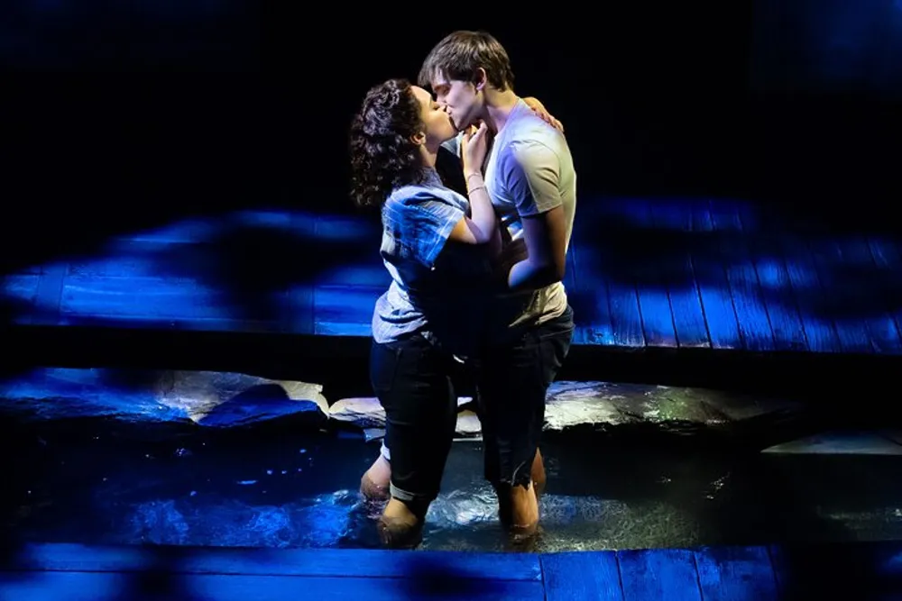 Two actors are sharing a romantic moment on stage with a kiss illuminated under blue stage lighting