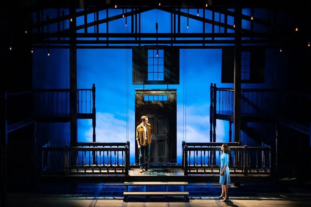 Two actors on a theatrical stage set featuring a two-level structure with a door at center stage under moody blue lighting evoke a dramatic or contemplative scene