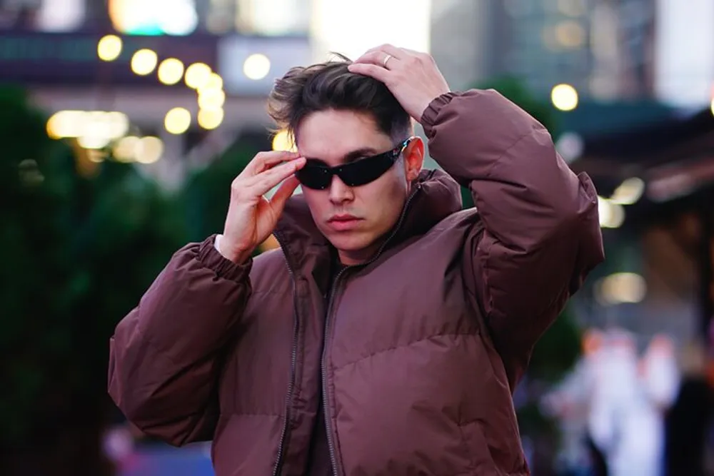 A person is adjusting their sunglasses and hair while wearing a brown jacket in an urban outdoor setting