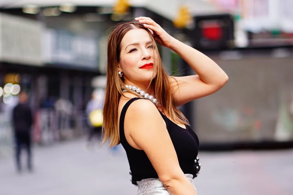 A person with red lipstick is striking a pose on an urban street wearing a black top and a large pearl necklace with a blurred city background