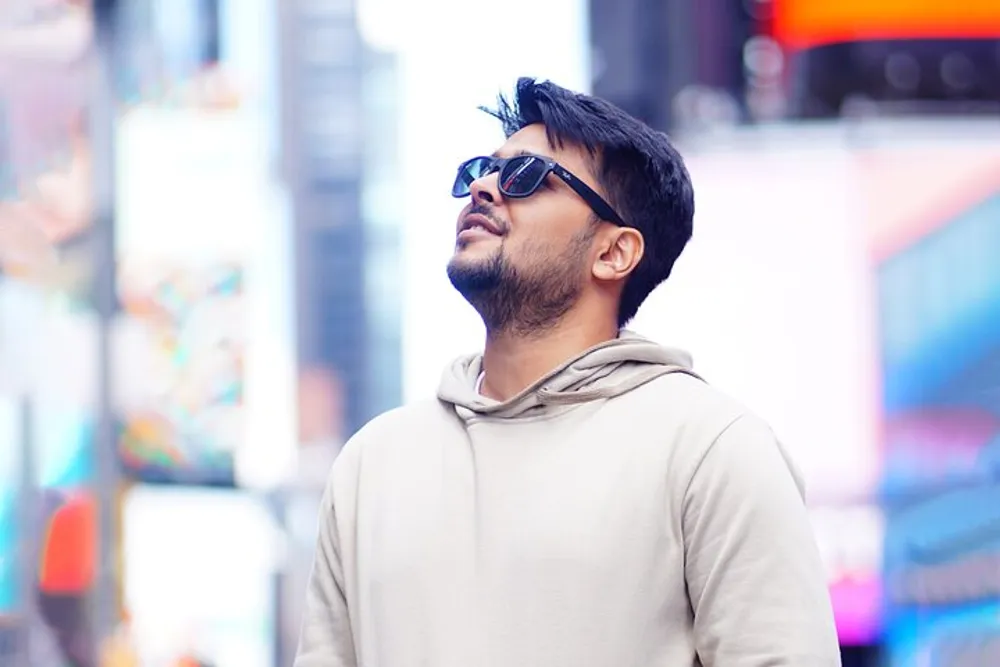 A man with sunglasses looks up thoughtfully against a blurred urban backdrop with colorful displays