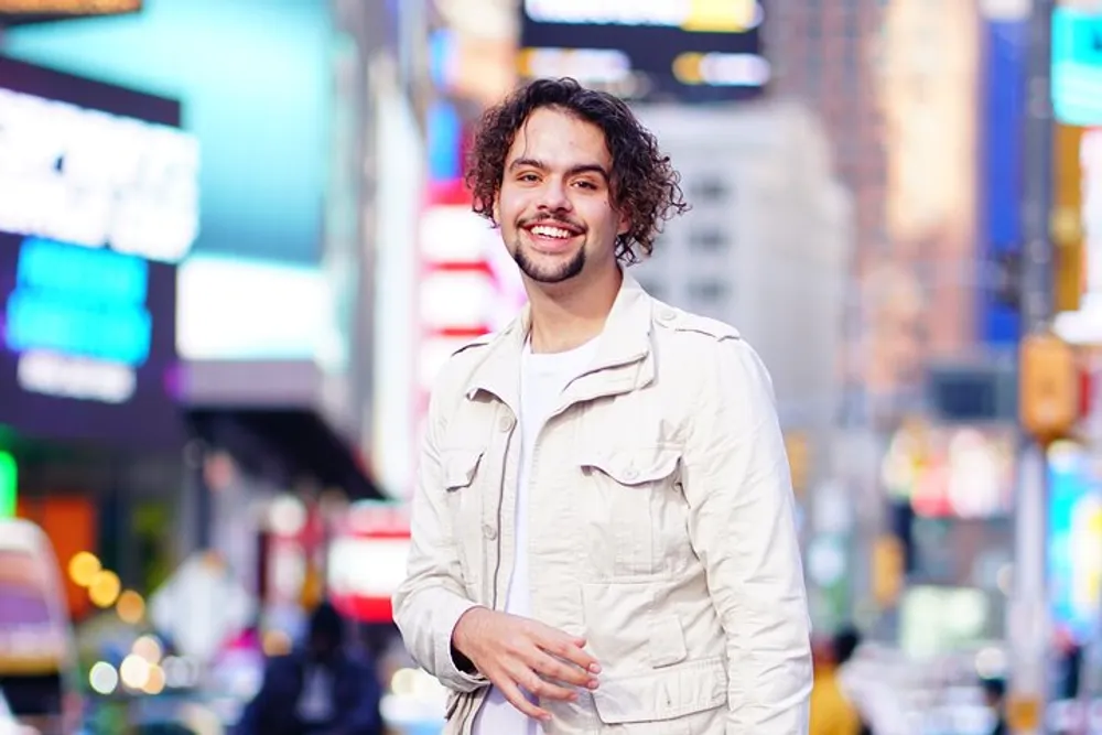 A person is smiling at the camera with a vibrant street scene possibly Times Square blurred in the background