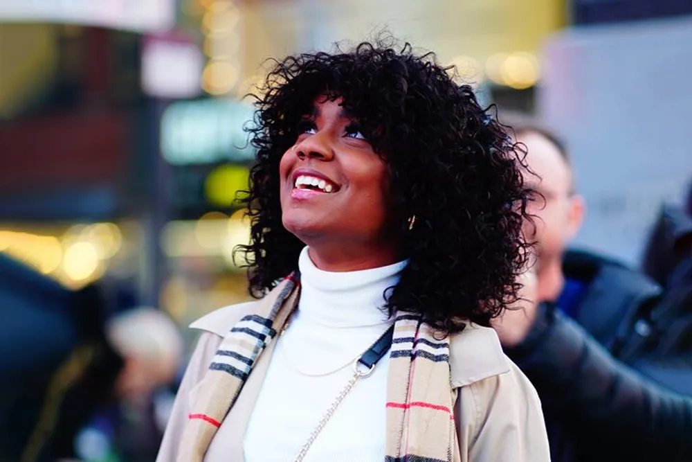 A joyful woman with curly black hair is looking upwards with a wide smile wearing a white turtleneck and a beige coat with a plaid scarf in an urban setting