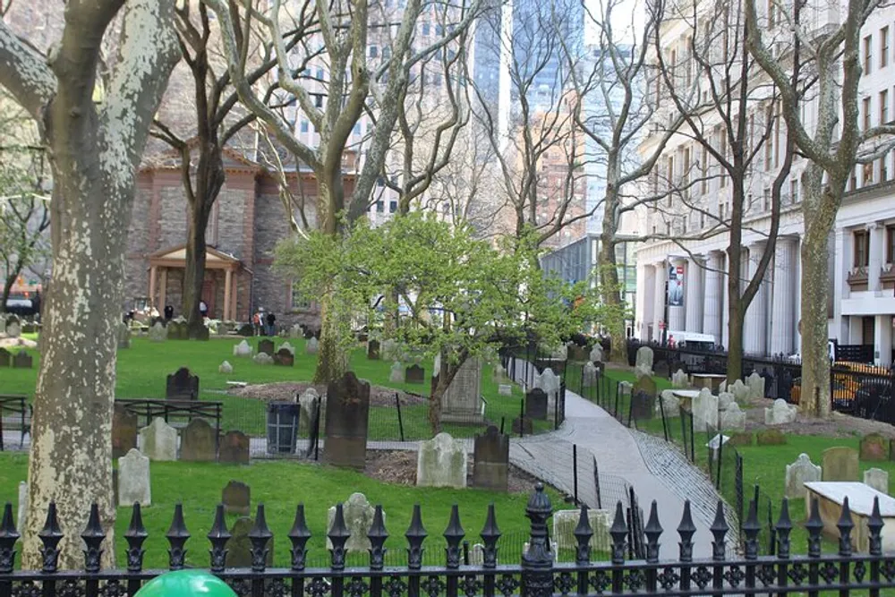 This image shows an old cemetery with weathered gravestones surrounded by leafless trees with a church in the background and modern buildings beyond creating a juxtaposition of the historical and contemporary in an urban setting