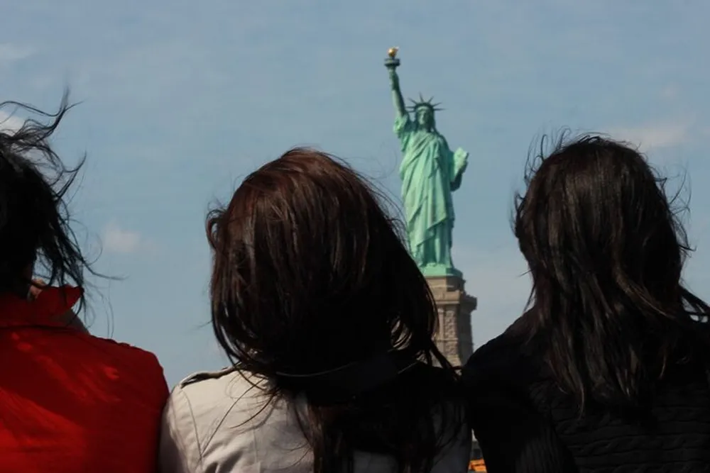 Two people are facing away from the camera looking towards the Statue of Liberty under a blue sky