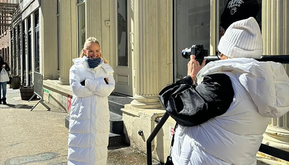 A person in a white coat is smiling while another person in white taking a photo captures the moment on a sunny day with building facades in the background