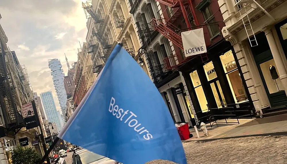 A blue flag with the words Best Tours dominates the foreground of an urban street scene lined with historic buildings