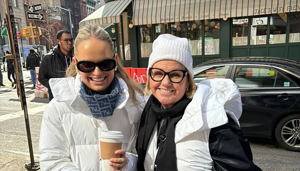 Two smiling women in winter clothing are holding coffee cups on a sunny city street