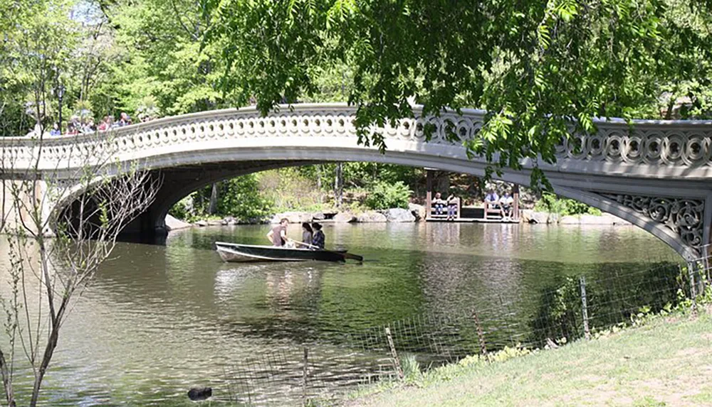 An ornate bridge spans over a tranquil body of water with people rowing a boat beneath and others enjoying the surroundings