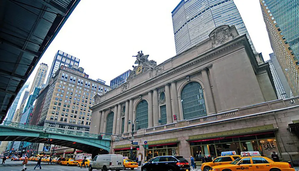 The image shows a bustling street view with iconic yellow taxis in front of a grand historic building with an elevated train track to the left in what appears to be a busy city center