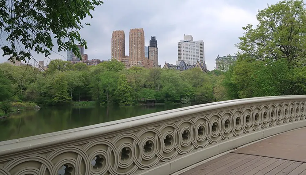 The image shows a scenic view from a bridge with a decorative railing overlooking serene water and lush greenery with city skyscrapers in the background under an overcast sky