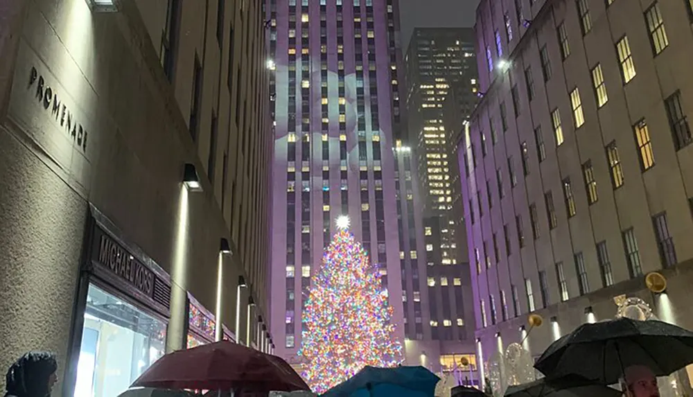 A vibrant Christmas tree illuminates a plaza surrounded by skyscrapers and people with umbrellas on a rainy evening