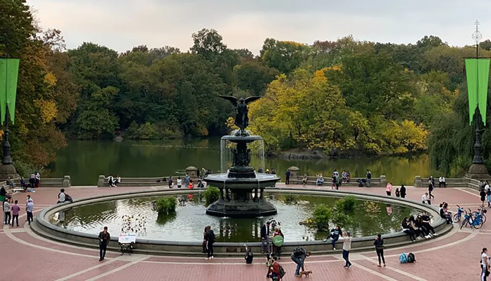 This image depicts a tranquil scene at a park with people enjoying leisure activities around a large ornate fountain with lush trees in the background showing early fall colors