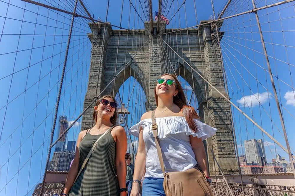 Two women are smiling and enjoying a sunny day on the Brooklyn Bridge with the New York City skyline in the background