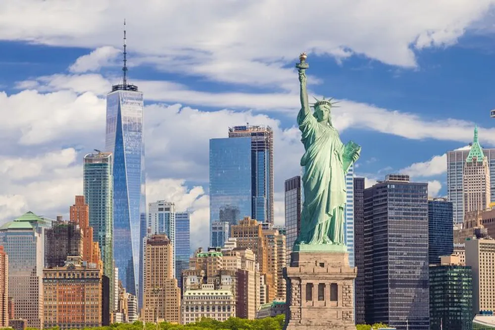 The image features the Statue of Liberty in the foreground with the New York City skyline including One World Trade Center in the background under a partly cloudy sky