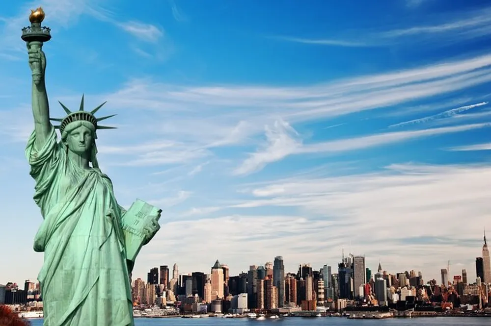 The Statue of Liberty stands prominently in the foreground with the New York City skyline stretching out in the background under a wide blue sky with wispy clouds