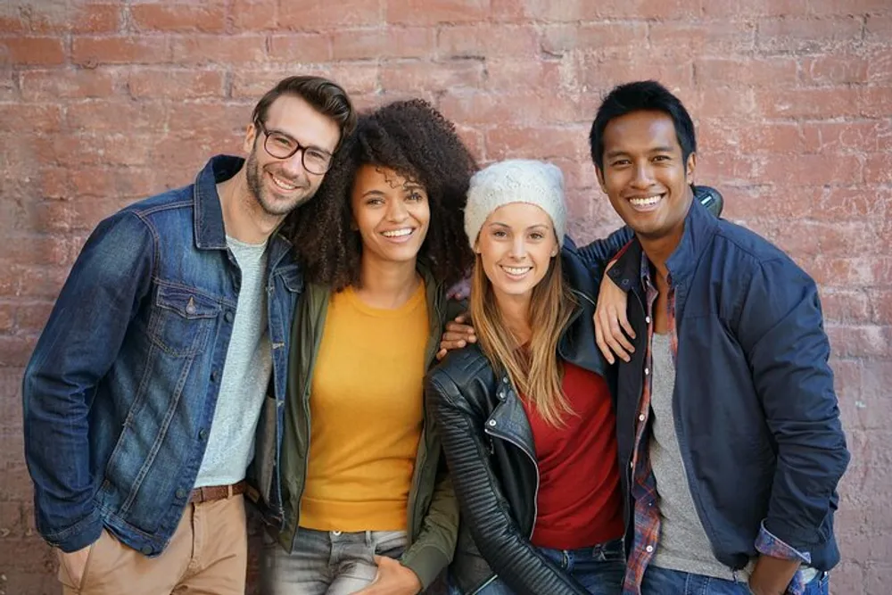 Four smiling friends are standing close together in casual clothing against a brick wall background