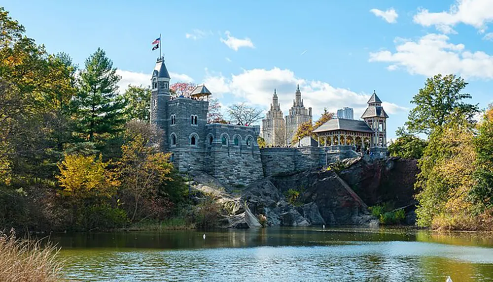 The image shows a picturesque castle-like building perched on a rocky outcrop overlooking a tranquil body of water set against a backdrop of autumn foliage and a clear blue sky