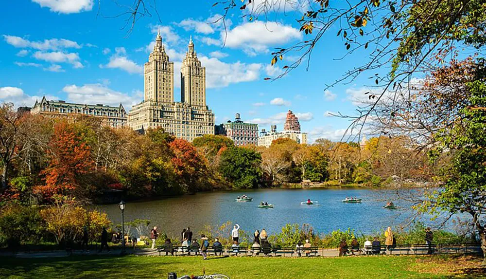 This image depicts a vibrant autumn scene with people enjoying leisurely activities by a lake in a park with towering buildings standing prominently in the background under a clear blue sky