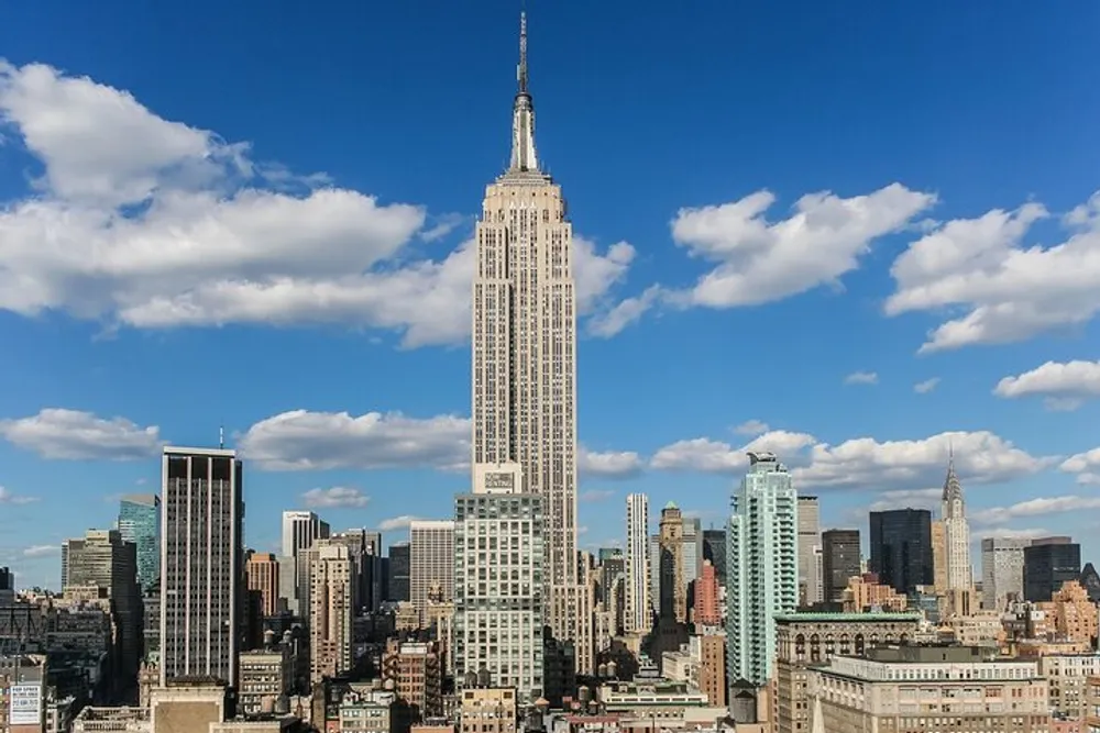 The Empire State Building towers above surrounding skyscrapers against a backdrop of a blue sky with fluffy clouds