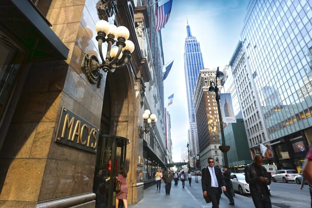 A bustling city street with people walking featuring the exterior of a building with the signage MACYS and a view of a tall skyscraper in the background under a clear sky