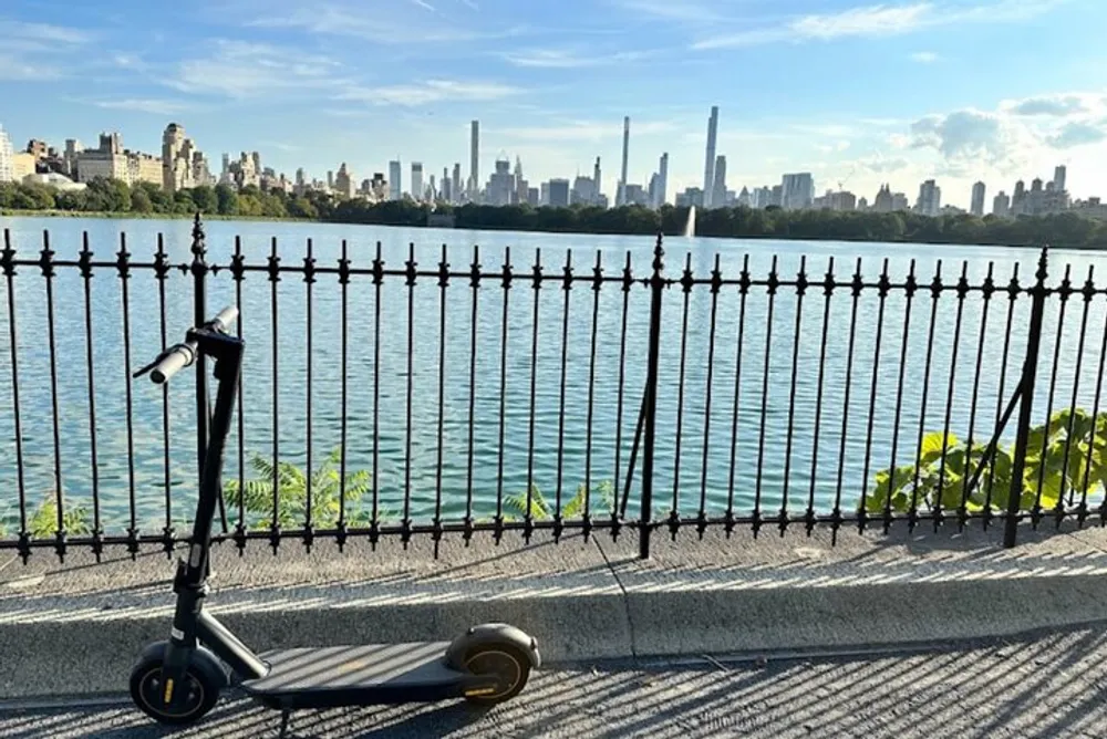 An electric scooter stands abandoned by an ornate metal fence with a scenic view of a city skyline across a body of water