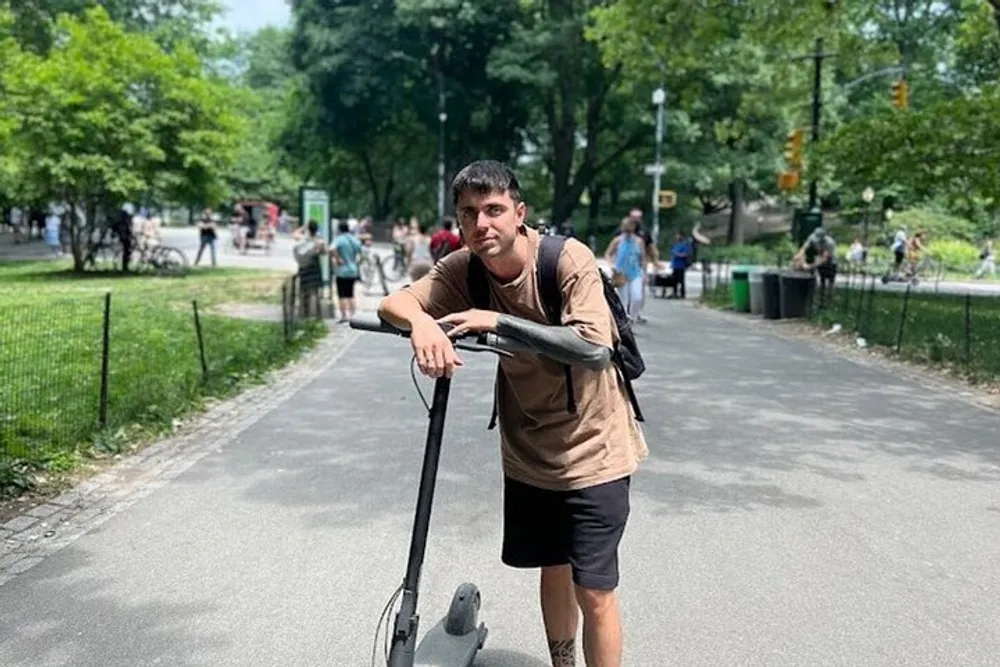 A person is leaning on an electric scooter posing for a photo in a park with trees and other people in the background