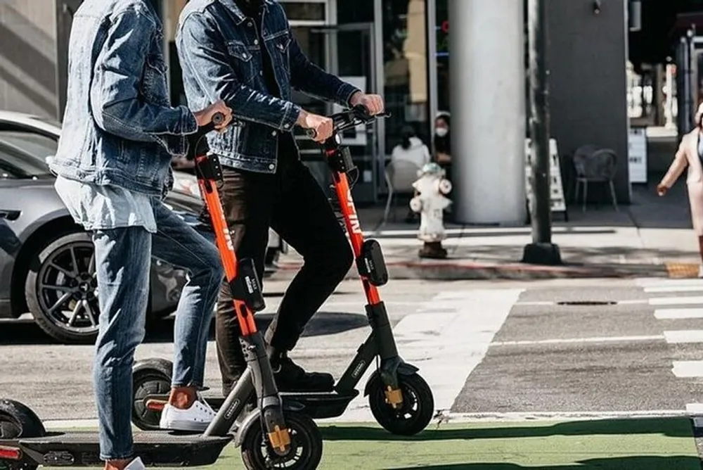 Two people are riding electric scooters across a street with one wearing a denim jacket and jeans while the other is dressed in black