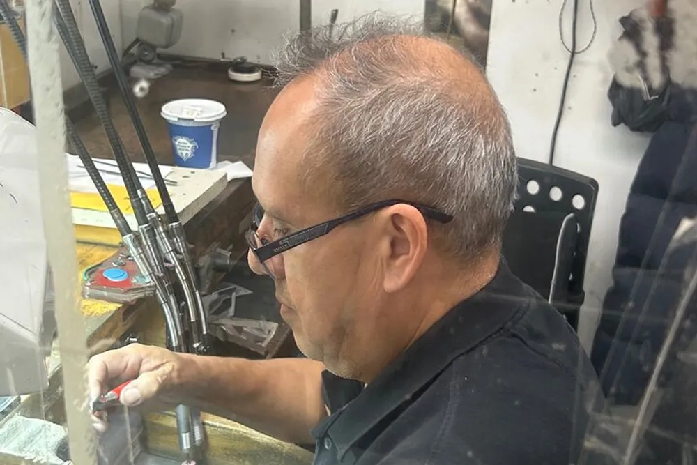 A person is seen working meticulously on a small object at a cluttered workbench possibly engaged in some form of craftsmanship or repair work